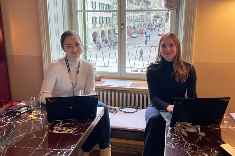 Zwei Frauen arbeiten mit Laptops an einem Tisch vor einem Fenster mit Stadtblick.