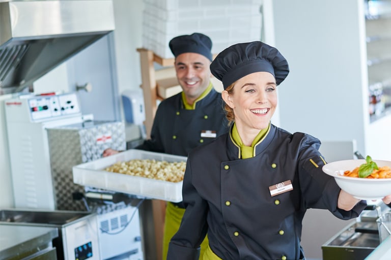 Zwei Mitarbeitende in der Küche lächeln und bereiten Essen zu, Frau steht im Vordergrund und reicht einem Gast einen Teller mit Essen.