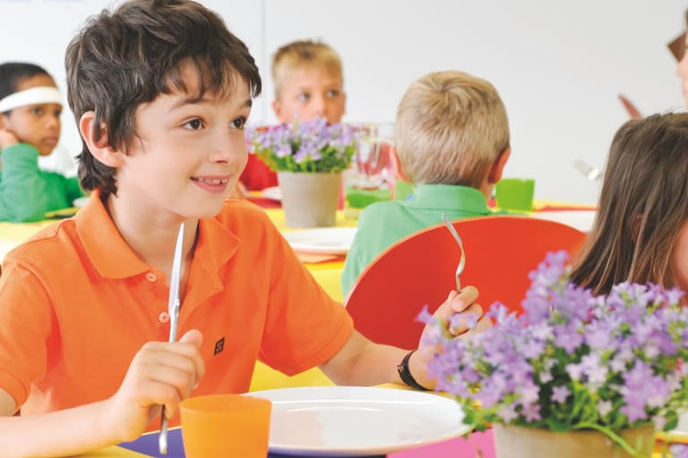 Junge in orange T-Shirt sitzt und lächelt am Tisch mit anderen Kindern in der Schulcafeteria.