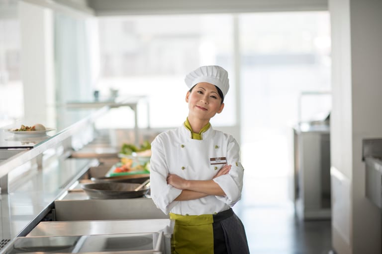 Eine Köchin steht in der hellen offenen Küche eines Restaurants mit verschränkten Armen und lächelt freundlich in die Kamera.