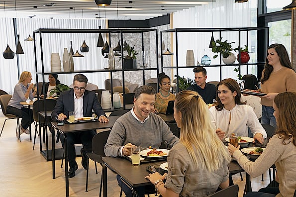 Gäste sitzen in einem gemütlichen und gut besuchten Mitarbeiterrestaurant ihres Unternehmens und geniessen ihr Mittagessen.