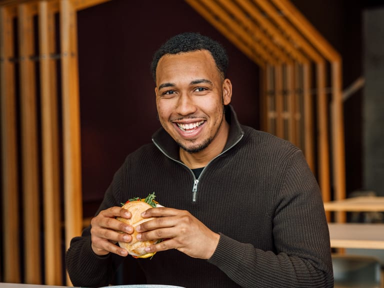 Lächelnder Mann hält einen Burger, sitzt in einem Restaurant.