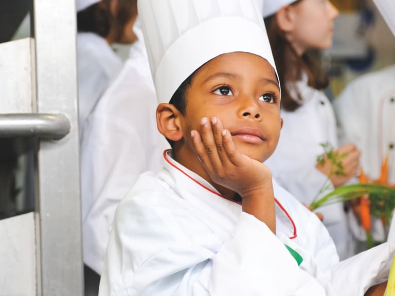 Kind mit einer weissen Kochuniform und Kochmütze sitzt in der Küche und denkt nach, andere Kinder sind im Hintergrund.