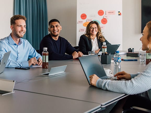 Menschen sitzen lächelnd in einem Besprechungsraum am Tisch mit Laptops.