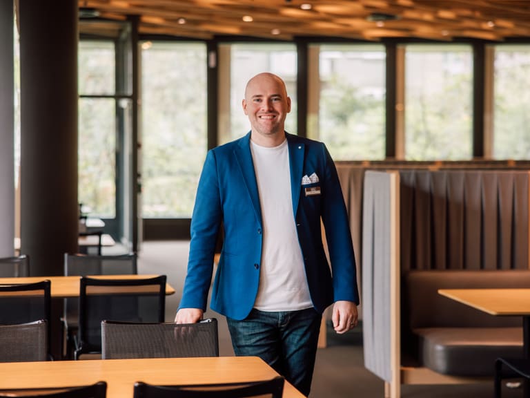 Mann in blauem Veston steht lächelnd in einem modernen Café.