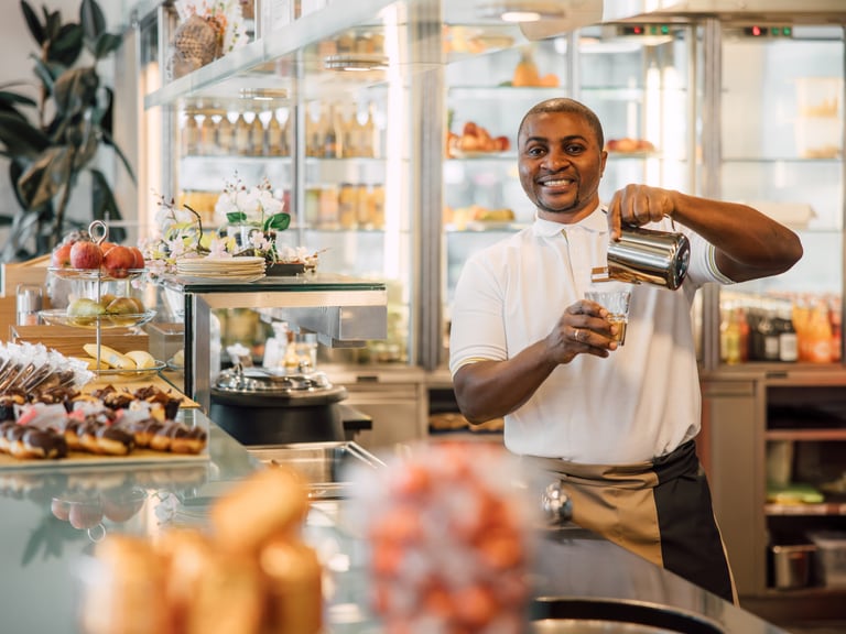 Mann steht hinter einer Kaffeebar, lacht in die Kamera und giesst gleichzeitig mit einer Kanne Milch in eine Kaffeetasse.