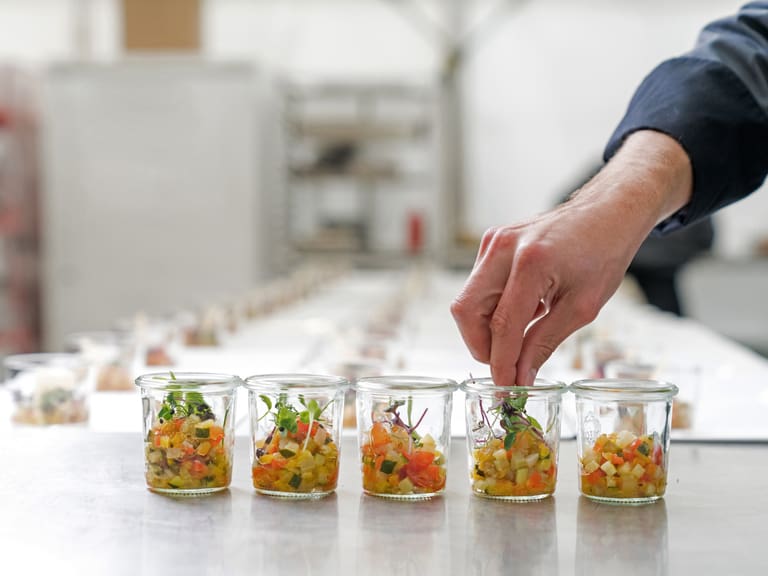 Person garniert kleine Gläser mit Gemüsesalat in einer Produktionsküche.