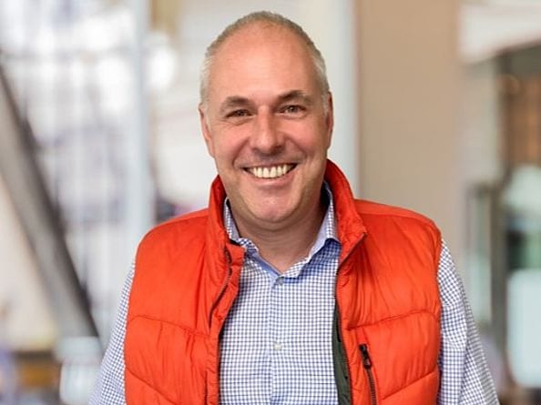 Mann mit lächelndem Gesicht trägt eine rote Weste und ein kariertes Hemd.