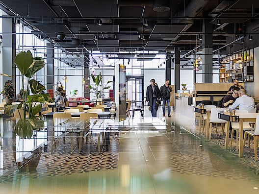 In einem modernen Mitarbeiterrestaurant mit Pflanzen und stylischem Mobiliar sitzen Gäste beim Mittagessen und laufen den Flur entlang.