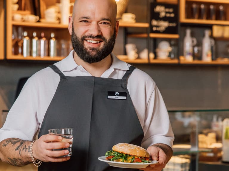 Mann in Schürze hält Teller und Glas in einem Café.