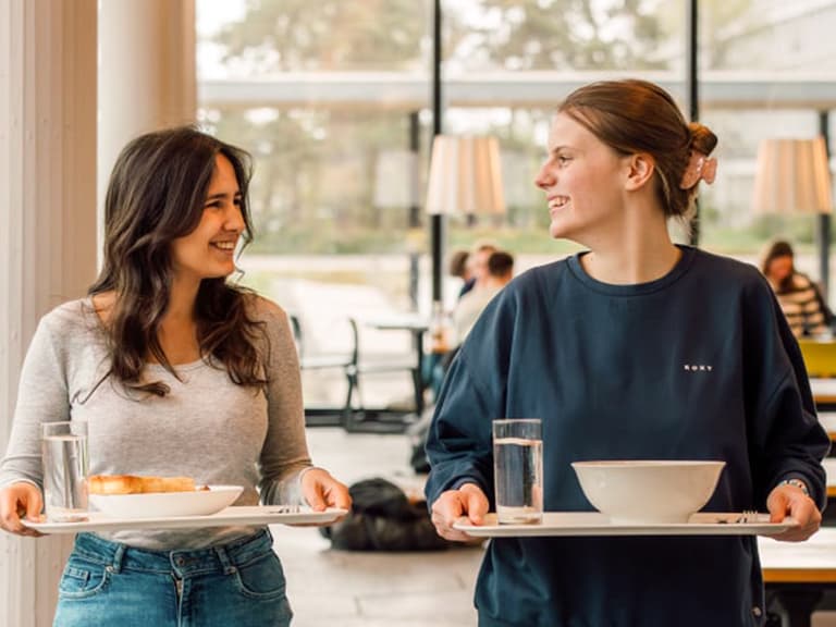 Zwei Frauen mit Tabletts im Essbereich, lächelnd.