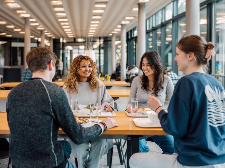 Gruppe von vier Personen sitzt und unterhält sich an einem Tisch in einer hellen Cafeteria