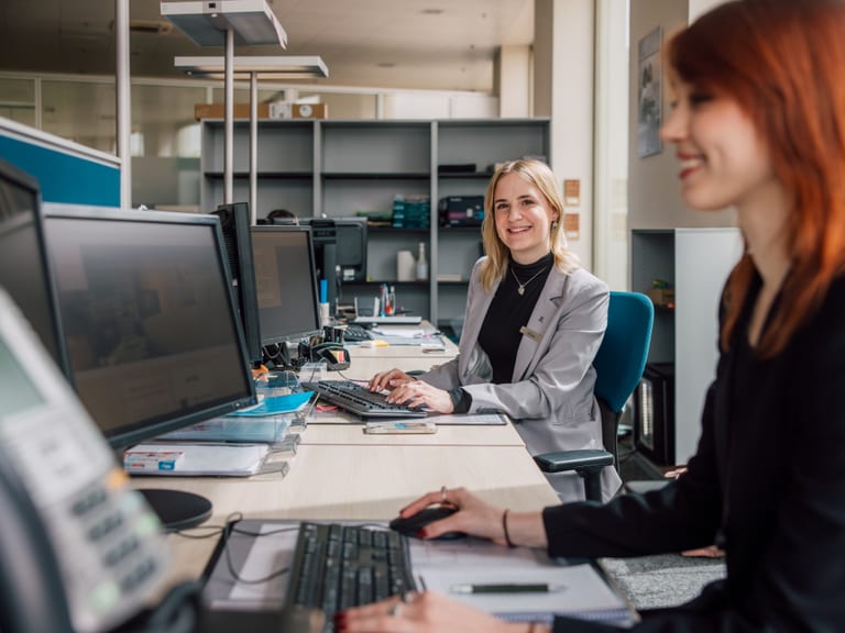 Zwei Frauen arbeiten lächelnd an Computern im Büro.