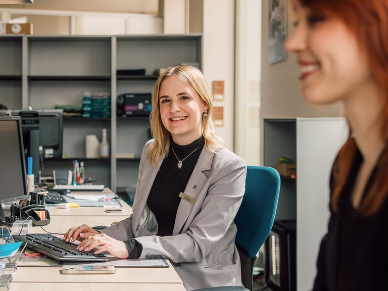Zwei Frauen an Büroarbeitsplätzen, eine lächelnd zur Kamera.