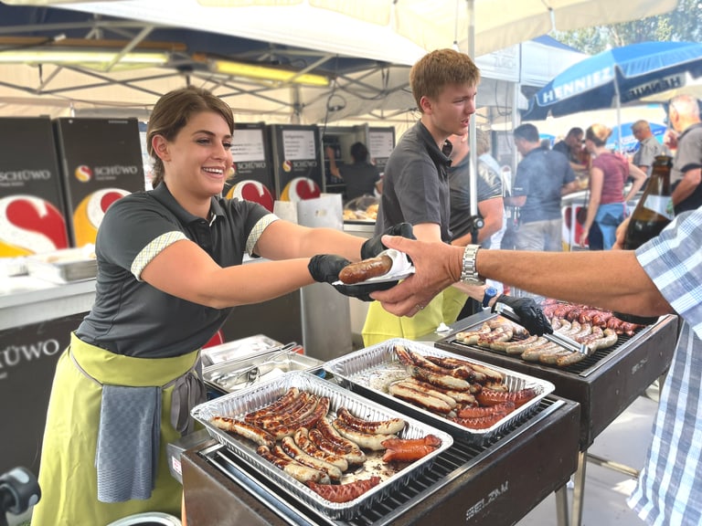 Mitarbeitede stehen an einem Grill bei einer Veranstaltung und geben frisch gebratene Würste an die Gäste heraus.