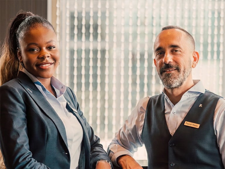 Zwei Hotelmitarbeiter in Uniform lächeln in die Kamera.