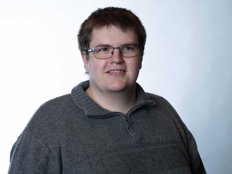 Person mit Brille in grauem Pullover, lächelnd vor hellem Hintergrund.