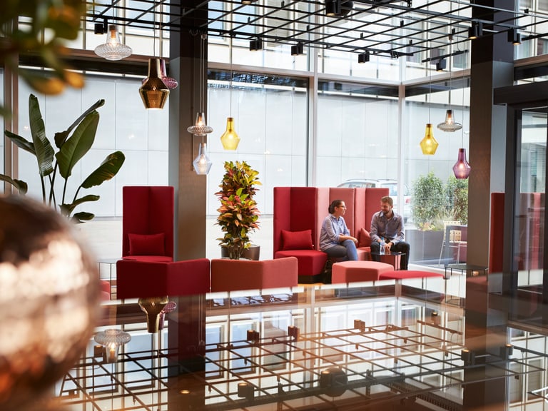 In einer modernen Lounge im Eingangsbereich eines Unternehmens unterhalten sich zwei Mitarbeitende.