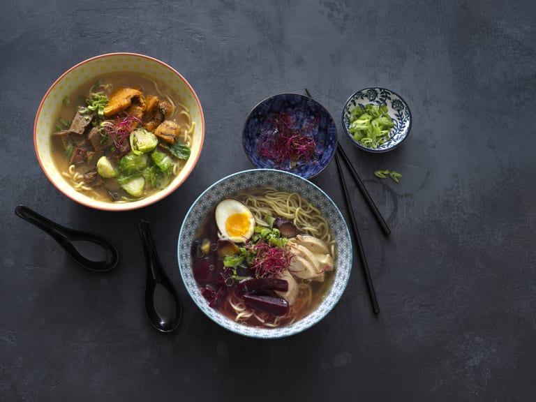 Zwei Schüsseln mit Ramen und Beilagen auf einem Tisch.