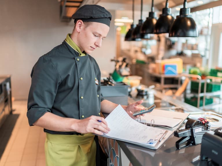 Ein Mitarbeiter in Kochuniform steht mit einer Checkliste an einem Counter.