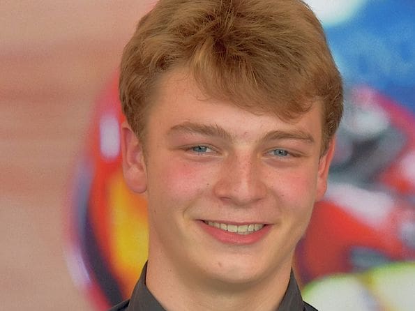 Lächelnder junger Mann mit grauem Hemd vor unscharfem Hintergrund.