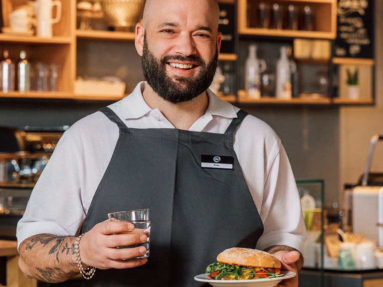 Lächelnder Mann in Schürze mit Sandwich und Getränk in Café.