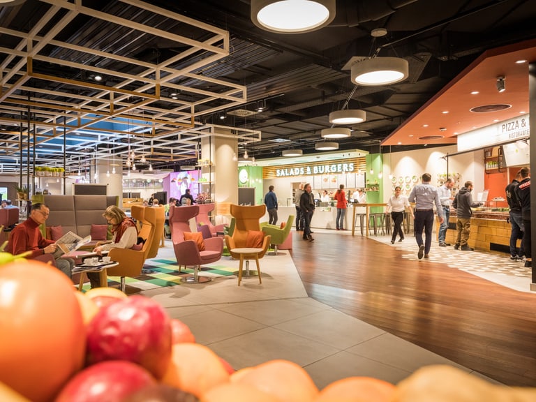 In einem grossen Foodcourt in einem Einkaufzentrum sitzen Personen in der gemütlichen Lounge oder an Tischen und geniessen Snacks, Kaffee, Kuchen oder Pizza, Pasta und Salate von den umliegenden Essensangeboten.