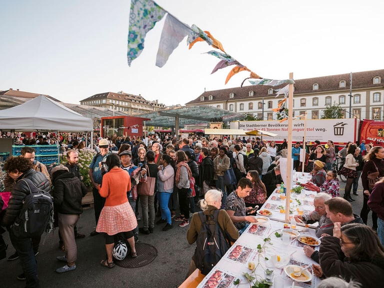 Versammlung von Menschen an einem Fest, im Vordergrund ein langer Tisch, an dem gegessen wird.