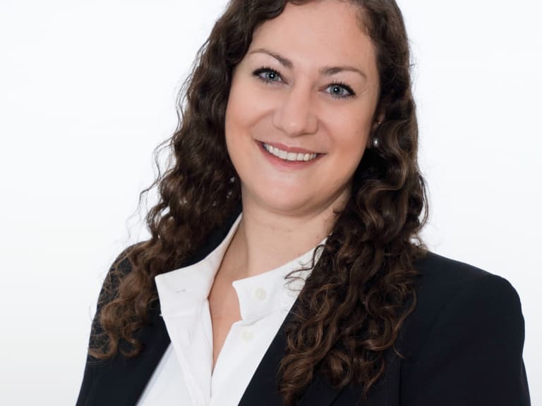 Lächelnde Frau mit langen, braunen Haaren vor hellem Hintergrund.