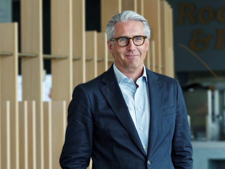 Mann mit grauem Haar und Brille in einem Anzug vor einem modernen Hintergrund.