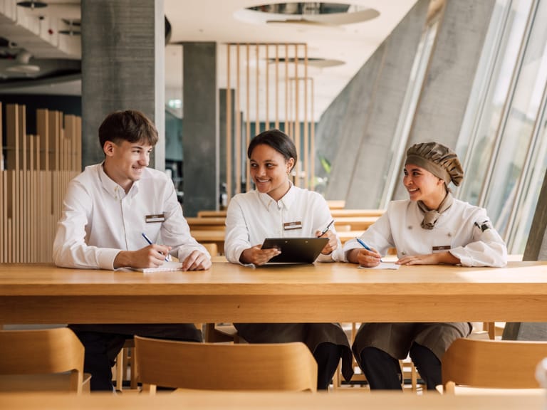 Drei Restaurantmitarbeiter in Uniform am Tisch sitzend, lächelnd.