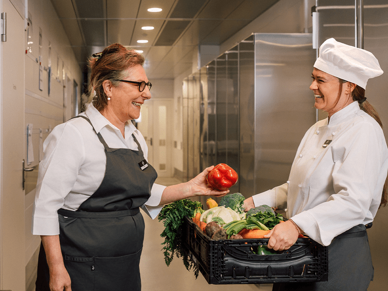 Zwei Köchinnen mit Gemüse in einer Küche lächelnd.