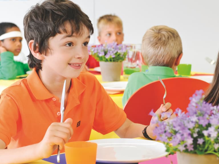 Junge in orange T-Shirt sitzt und lächelt am Tisch mit anderen Kindern in der Schulcafeteria.
