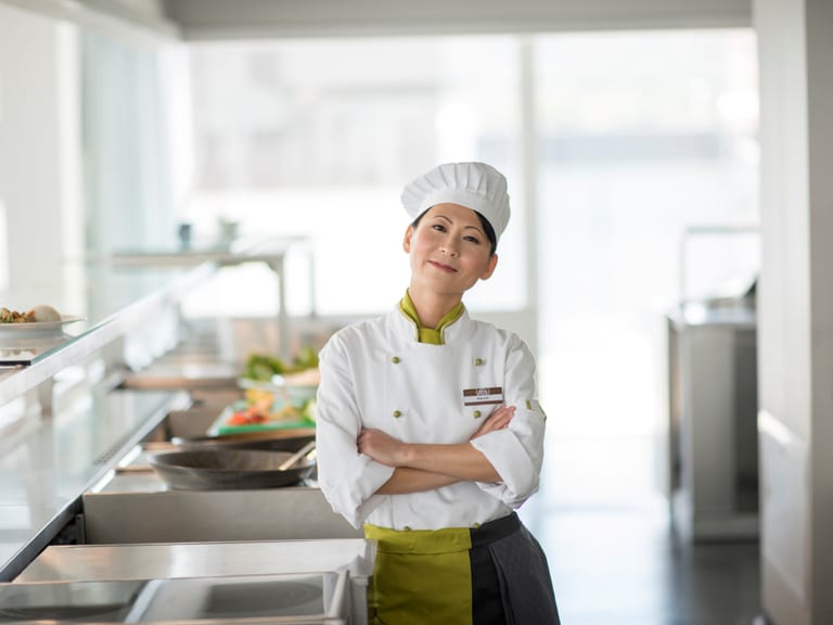 Eine Köchin steht in der hellen offenen Küche eines Restaurants mit verschränkten Armen und lächelt freundlich in die Kamera.