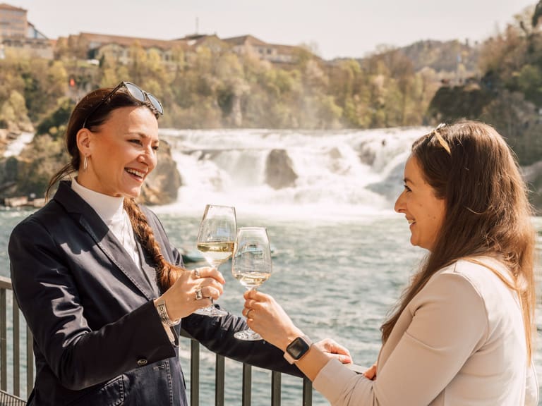 Zwei Frauen stossen mit Wein an vor Wasserfallkulisse.