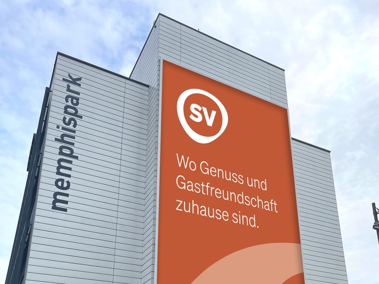 Gebäude mit grossem orangem Banner, 'SV' Logo und Text in deutscher Sprache.
