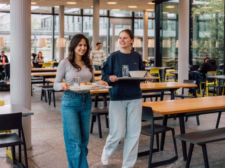 Zwei Personen mit Tabletts in einer Kantine gehend.