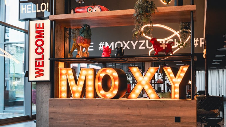 Empfangsbereich mit leuchtendem MOXY-Schild und Dekorationen.
