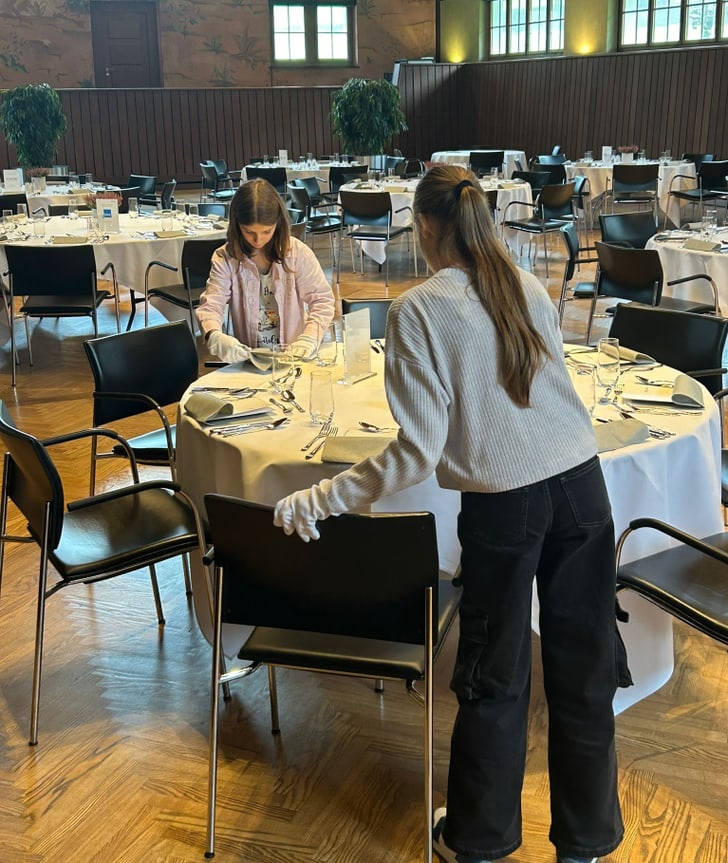 Zwei Personen decken Tisch in einem Restaurant mit weissen Tischdecken.",